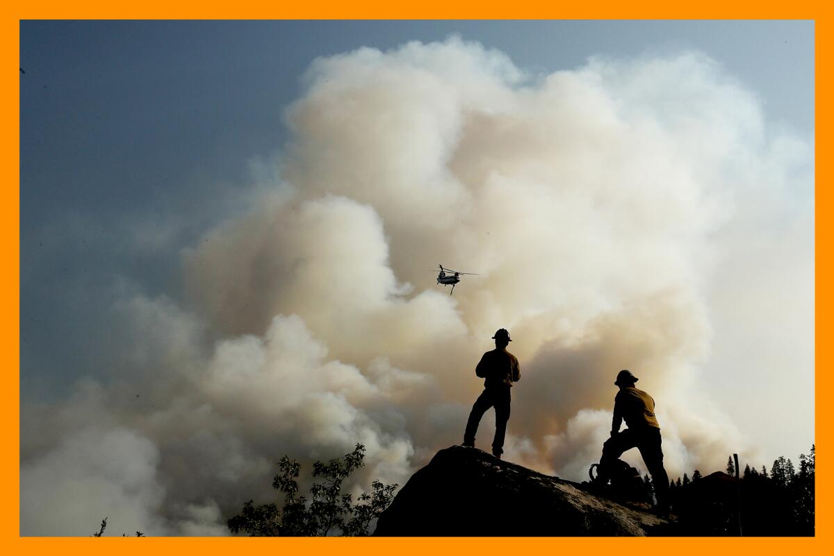 Firefighters stand on a rock watching a helicopter silhouetted against wildfire smoke