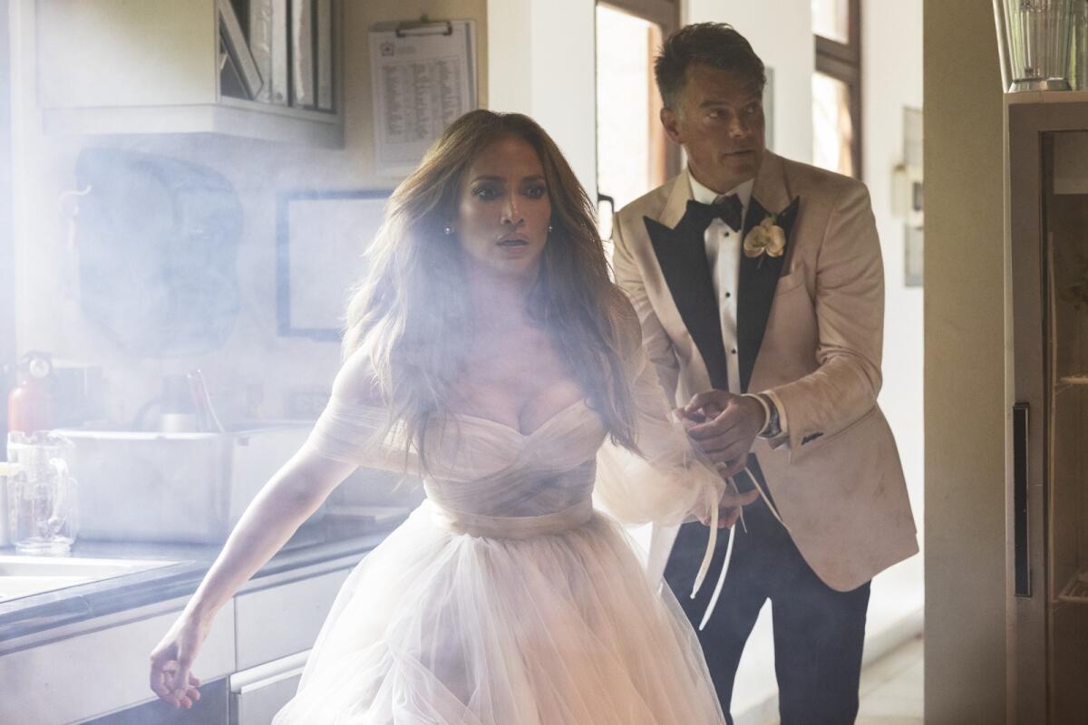 A long-haired woman in a low-cut dress stands in front of a man in a tuxedo in the movie "Shotgun Wedding."