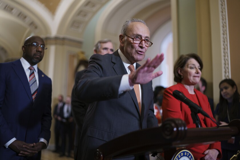 A man in a suit and glasses, flanked by a man and a woman, gestures before a microphone.
