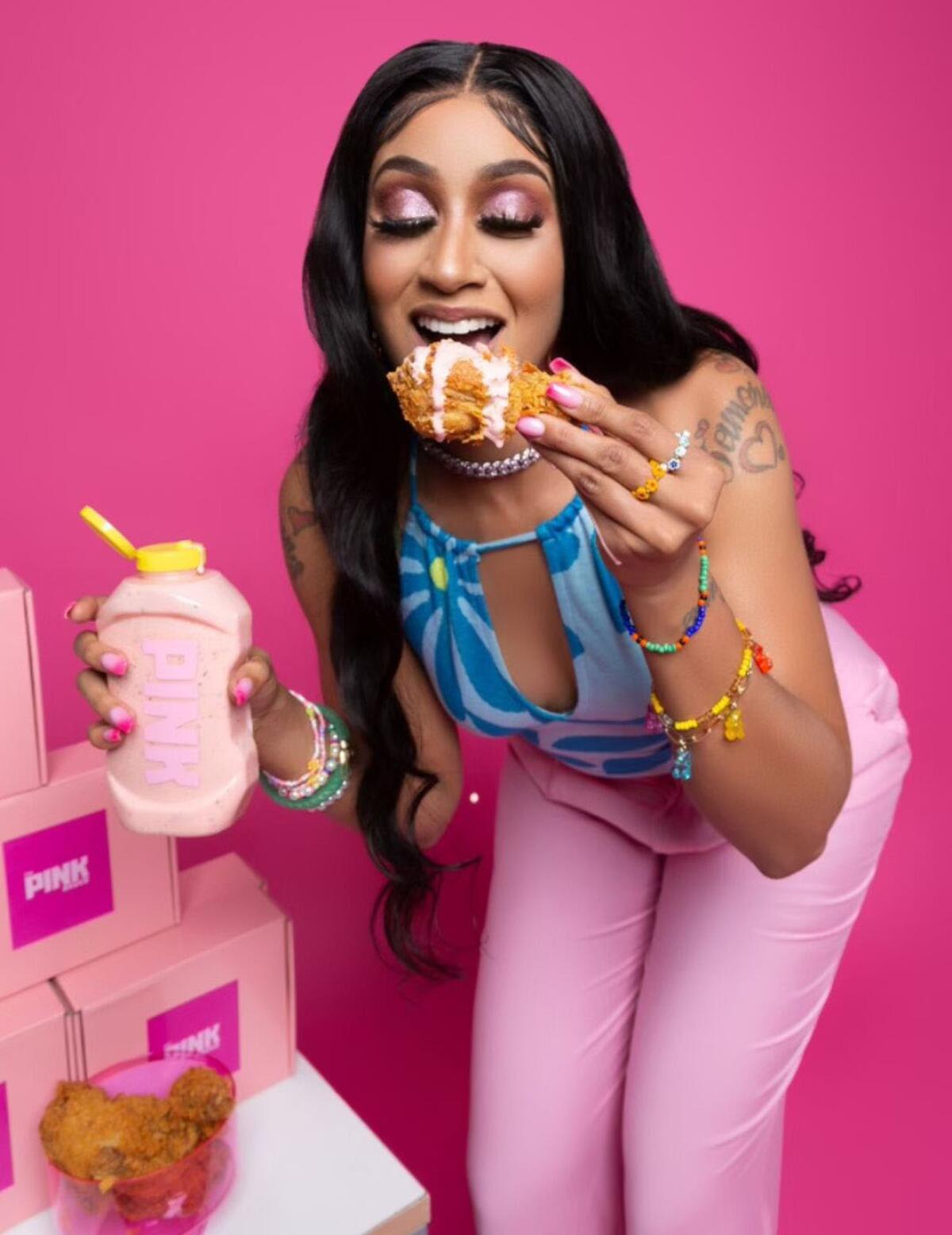 A woman holding a bottle of pink-colored sauce bites into fried chicken dipped in the condiment.