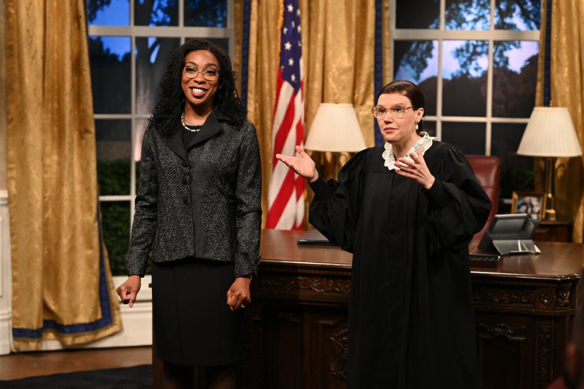A woman wearing a black blazer stands next to a woman wearing black robes on a set designed to resemble the Oval Office