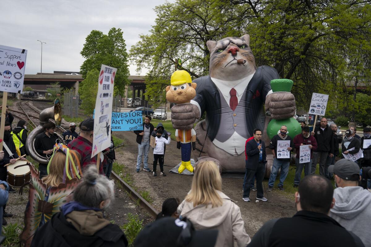 A man speaks to a small crowd outdoors while standing in front of a balloon depicting a "fat cat."