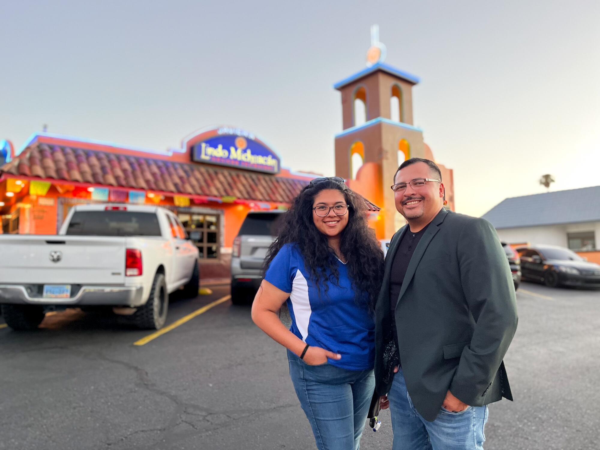 A woman with dark hair, in a blue shirt, left, and a man in a gray jacket, smile while standing outside a restaurant