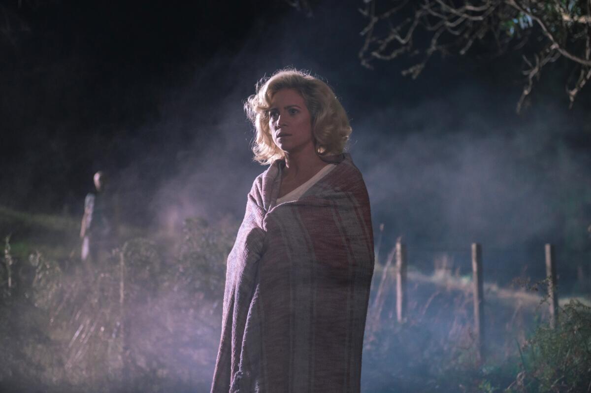 A woman stands in a field holding a blanket around her shoulders.