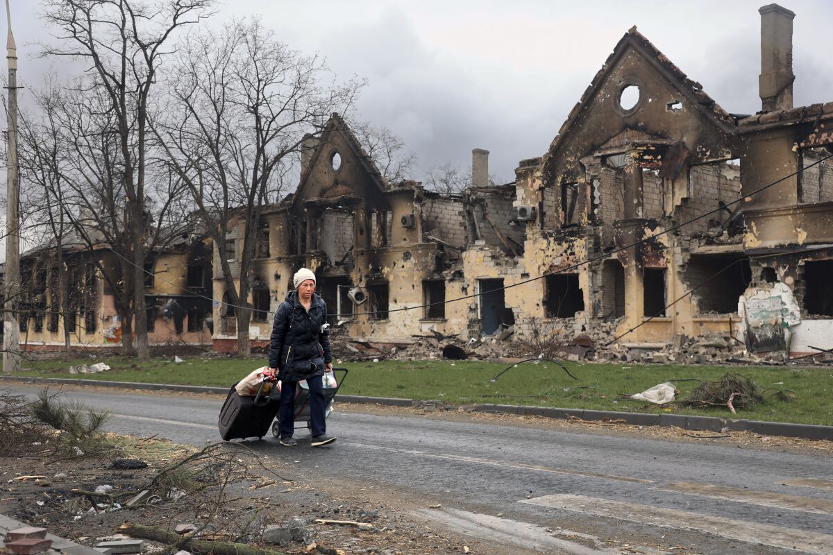 A woman pulls her luggage along a road, past charred and blown-out houses