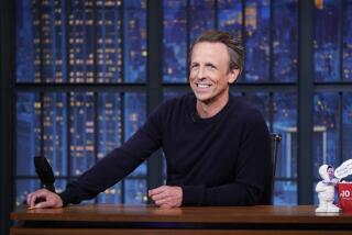 Seth Meyers smiles while sitting behind his desk on his late-night TV set.