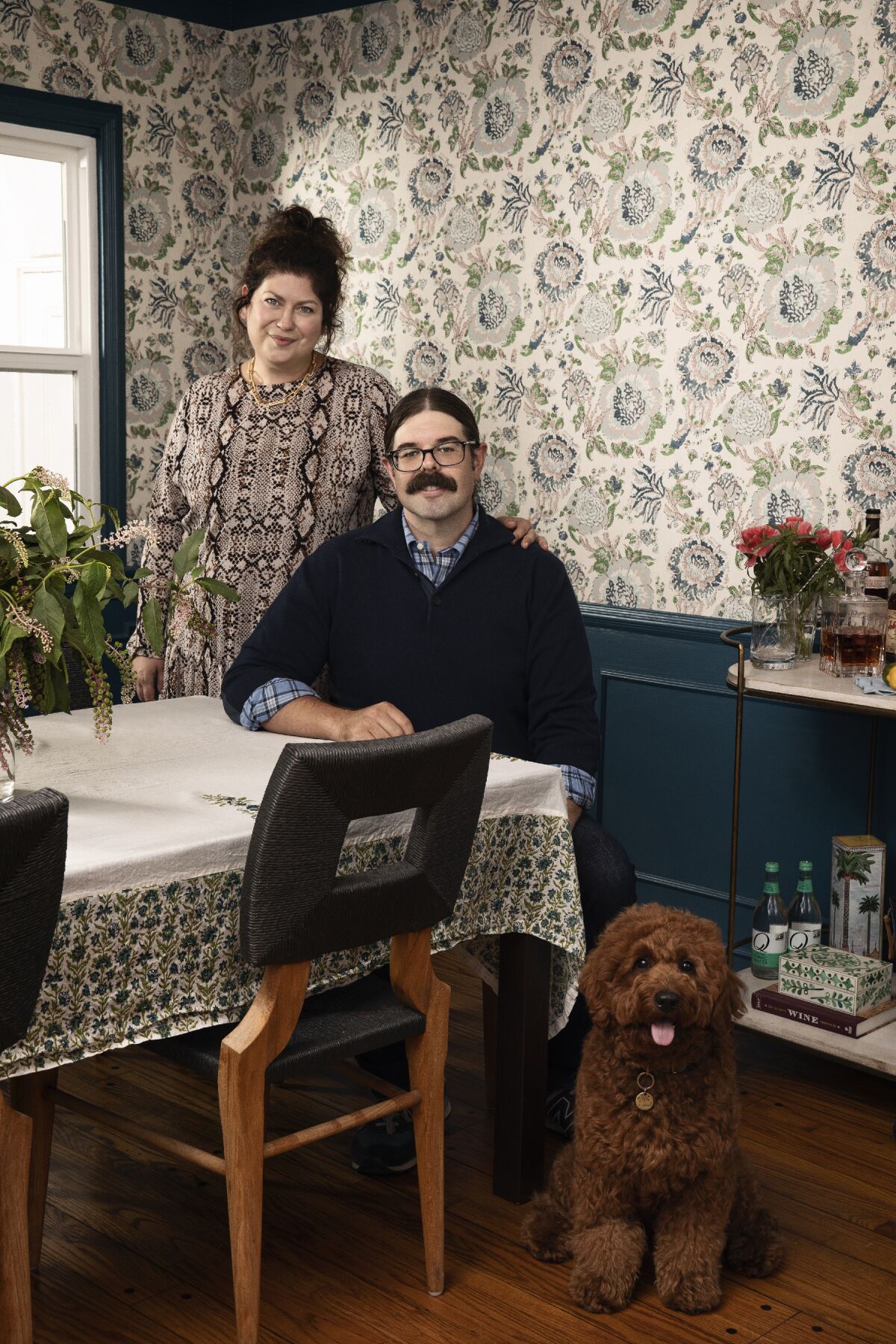A man and woman and their dog pose in their decorated dining room.
