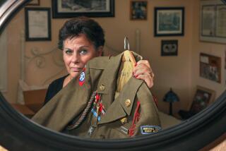 Portrait of Ginni Field, reflected in a mirror, holding the U.S. Army uniform of her late father Richard "Dick" Field. On the wall behind her are some of the many awards and honors from his combat experiences with the 551st Parachute Infantry Battalion in Operation Dragoon and the Battle of the Bulge, among others.