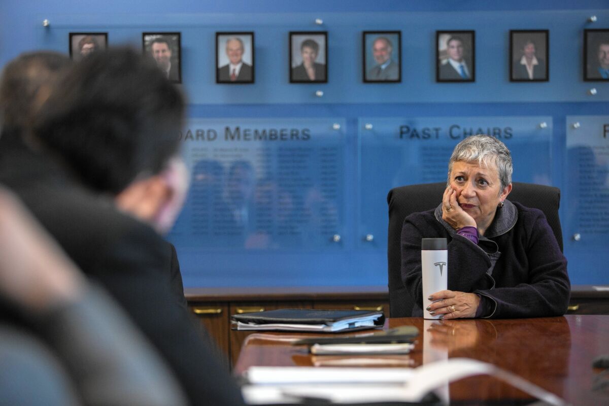 Mary Nichols sits at a table in a room during a meeting with framed photographs of people behind her.
