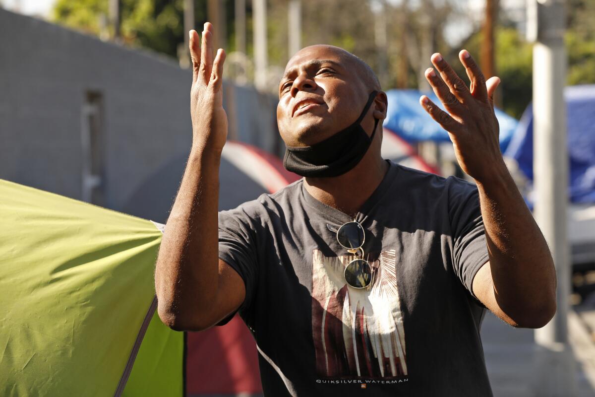 A man with a mask around his chin raises both hands to the sky in front of a row of tents on the sidewalk