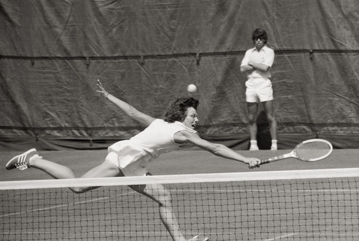 Wearing all white, Billie Jean King runs and reaches with a tennis racket to hit a shot at the net while a ball boy watches.