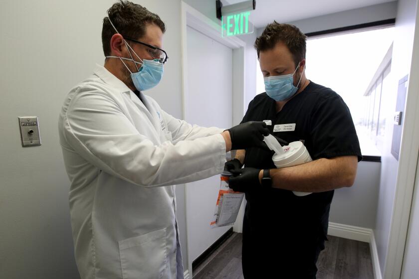 With his office manager Daniel Zike giving a hand, right, Dr. Matthew Abinante, left , prepares to take drive-up sample swabs for the novel coronavirus COVID-19 from patients while they remain in their vehicles, outside the offices of Elevated Health in Huntington Beach on Thursday, March 19, 2020.