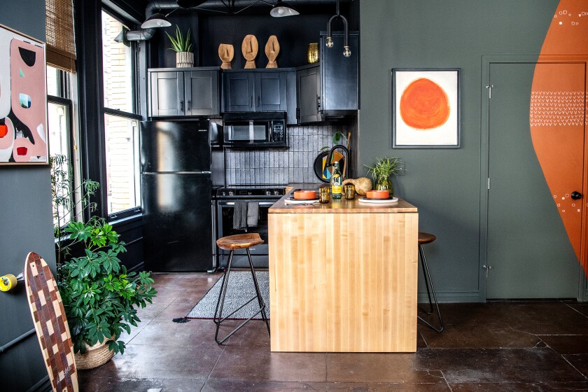 A kitchen with a refrigerator, oven and microwave and a wooden countertop. To the left side are a longboard and a plant.  