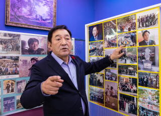 A man with dark hair, in a dark jacket, has one hand on one of two large framed displays of pictures on a wall.