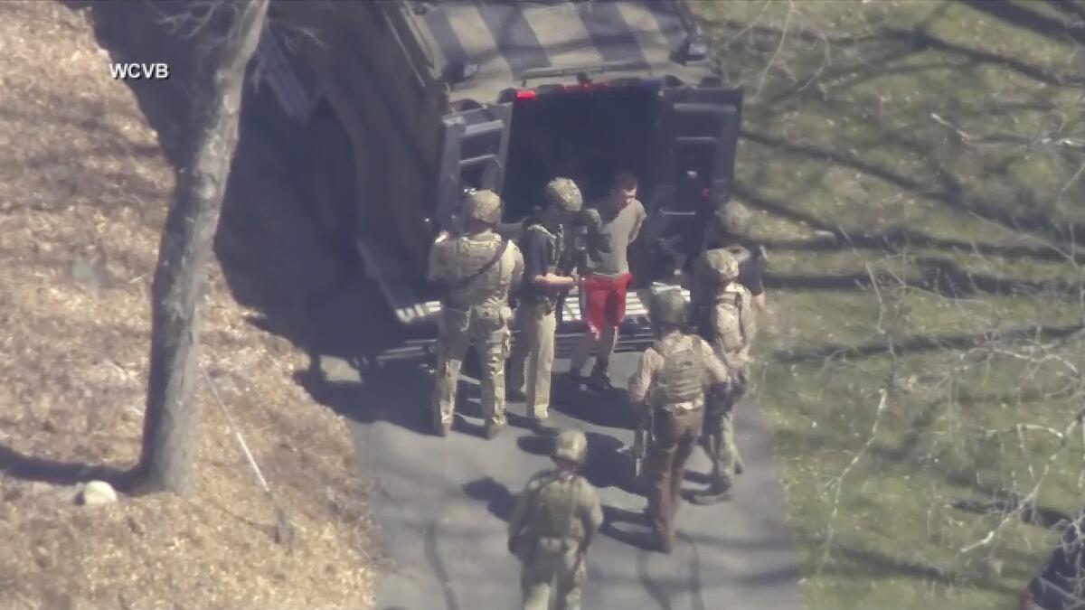 Officers in tactical gear surround a man in sportswear near the open back doors of a custody van.  