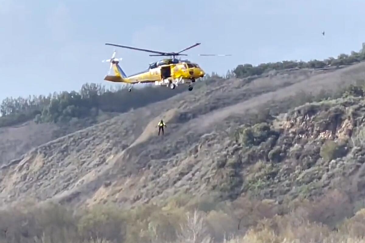 A person is seen suspended from a yellow helicopter against a hillside backdrop.
