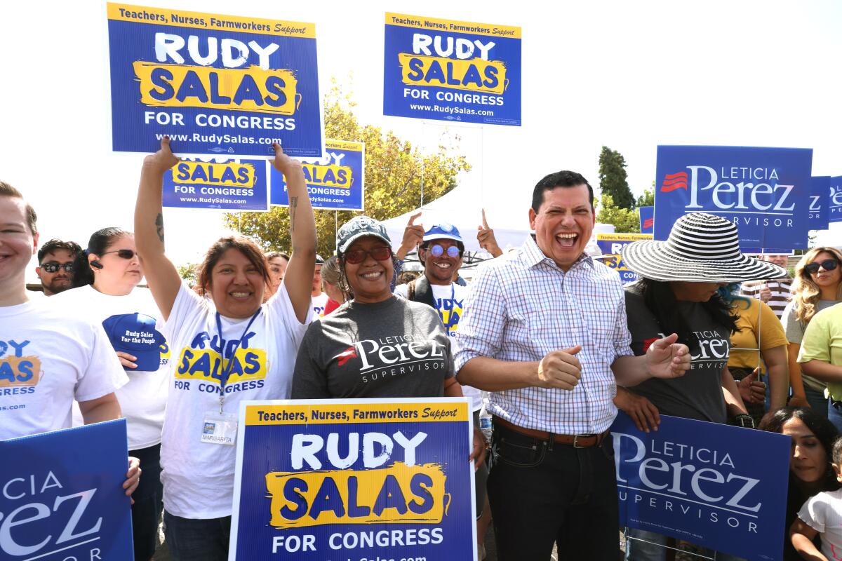 Rudy Salas, grinning widely, stands surrounded by supporters holding signs promoting his run for Congress. 