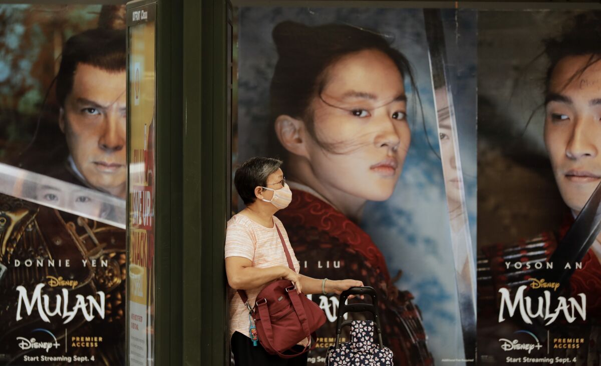Movie posters serve as a backdrop for a mask-wearing passenger waiting for her bus on Broadway in Chinatown.