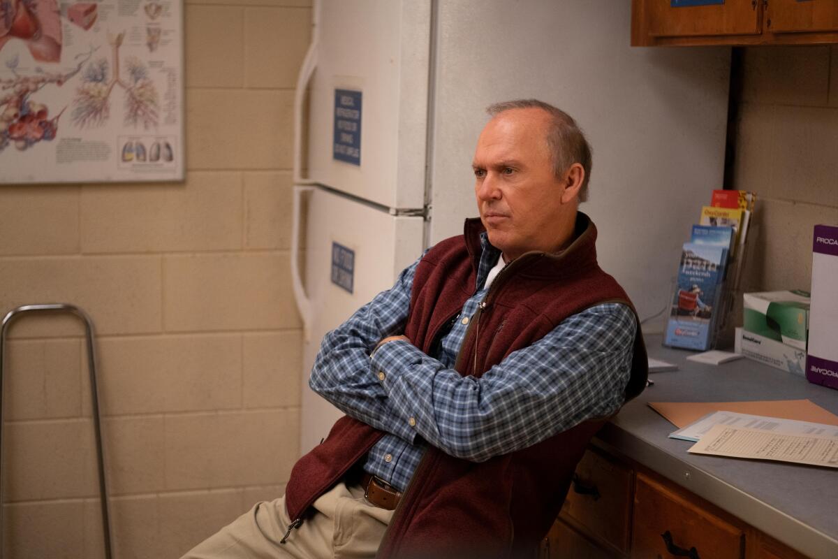 A man sits with his arms crossed, leaning against a counter next to a refrigerator. 