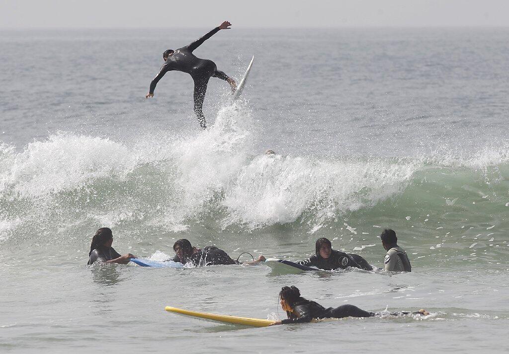 Participants of the Save Our Youth’s Surf Days program paddle in the water as a surfer flies over a wave in the background in west Newport on Thursday.