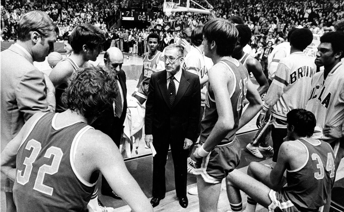 In a black-and-white photo, coach John Wooden wears a suit and talks seriously to players on the court.