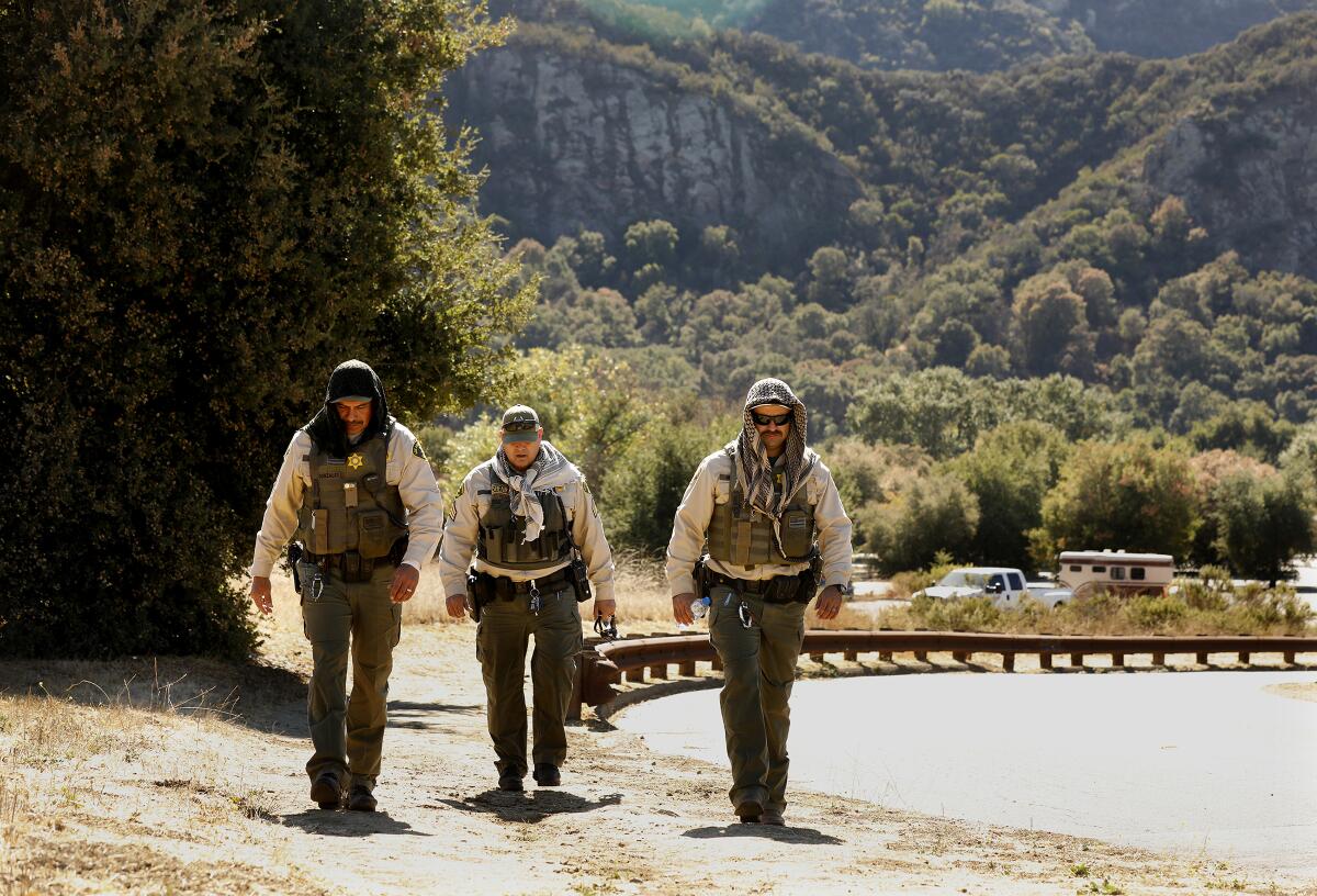 Three men wearing law enforcement uniforms and scarves walk in the sunshine along a road leading uphill from a forested area