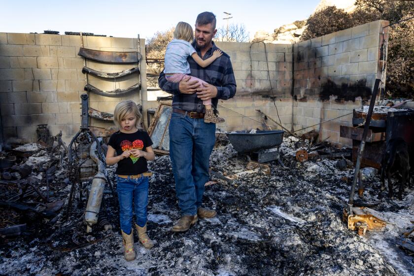 AGUANGA, CA - NOVEMBER 22, 2023: Single dad Cody McCormick with his two daughters Allison, 4, and Kayla,6, walk through parts of their destroyed garage which was reduced to ashes along with they home after the October Highland fire swept through on November 22, 2023 in Aguanga, California. He and the girls are staying in a borrowed trailer on the adjacent property. (Gina Ferazzi / Los Angeles Times)