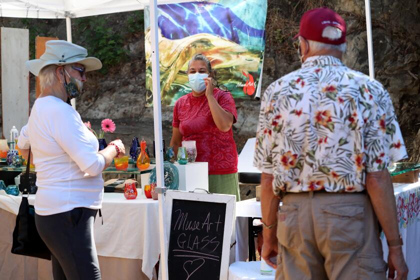 Glassblower Mary AnnGuerra talks to customers at the Sawdust Festival on opening day, in Laguna Beach on Saturday, Sept. 19, 2020. Vendors have artwork, photos, paintings, ceramics, glass, garments jewelry and a variety of other items for sale. The festival is implementing safeguards to ensure visitors safety by requiring face masks and social distancing. There are hand sanitizing stations and, according to the website, employees continually clean public areas, dining area and high touch areas along with restrooms, There is a capacity of 250 people at a time and vendors have also been limited to give allow more space for social distancing.