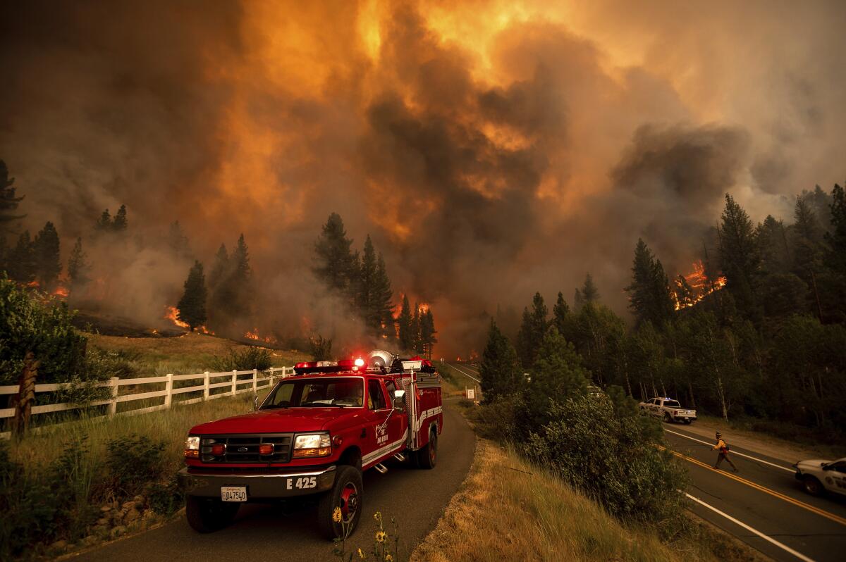 A fire truck is parked by a road as flames roar behind it