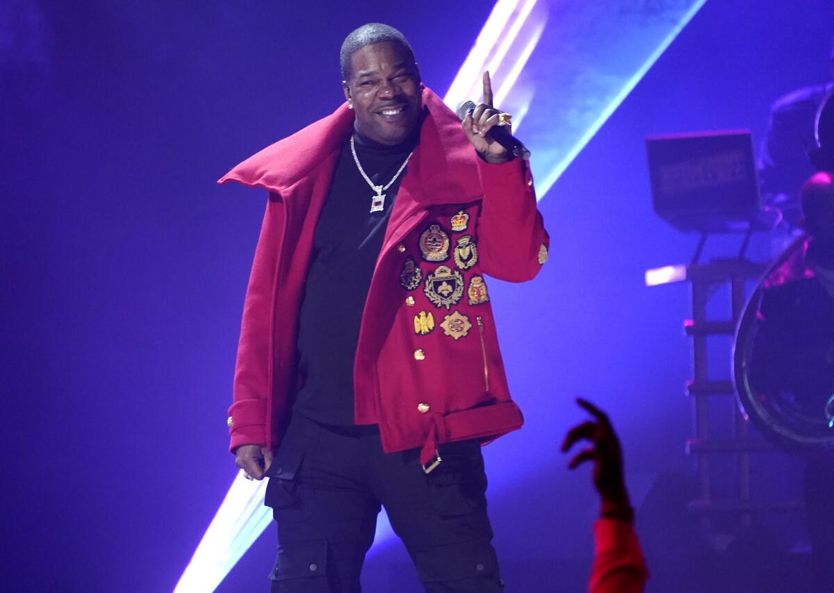 A man in a red jacket and silver necklace standing on a stage, smiling and pointing a finger upward