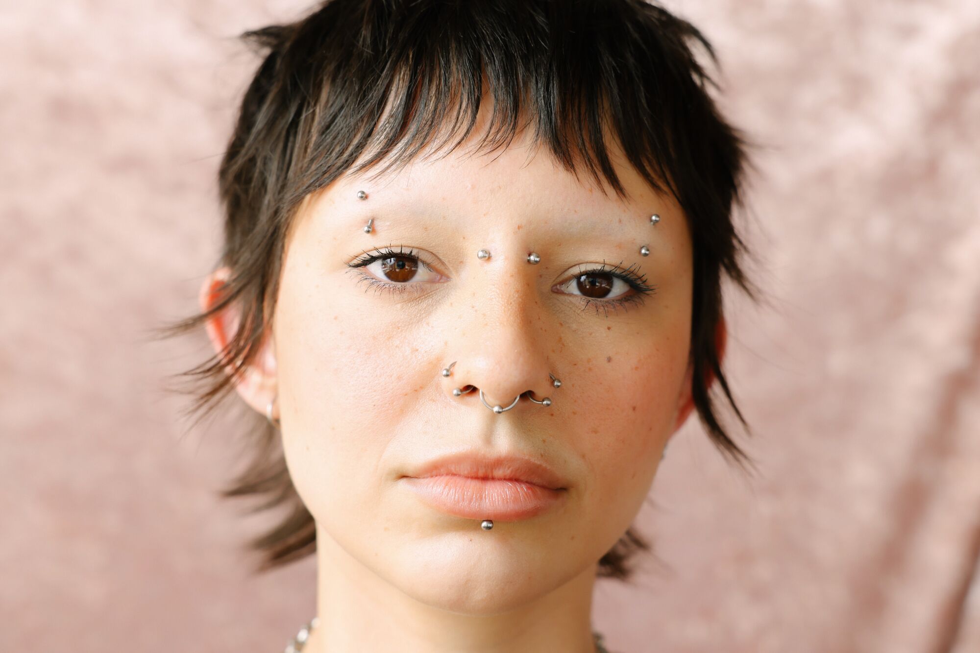 A person with facial piercings short hair feathered around the face looks into the lens.