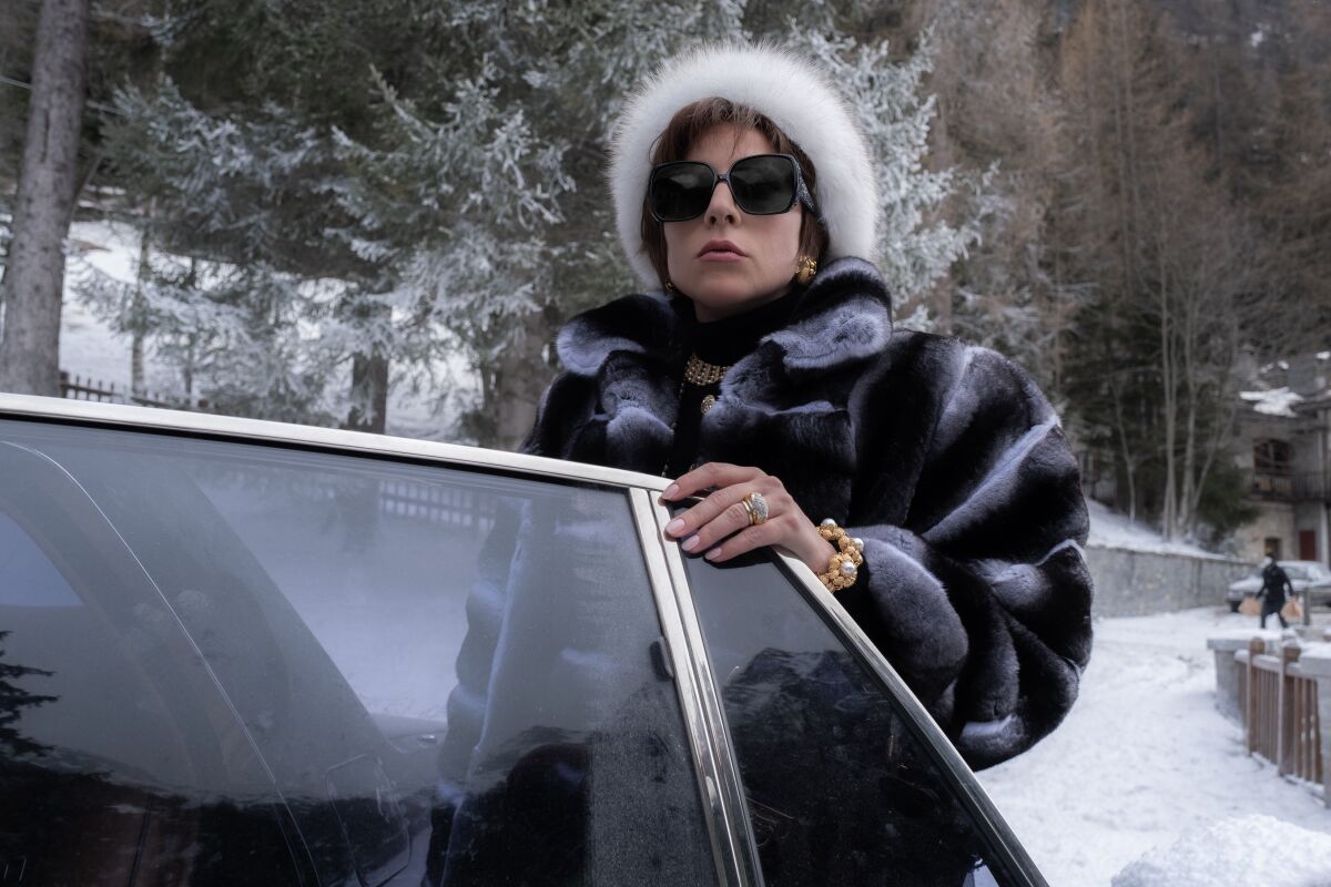 A woman in a fur hat, fur coat and sunglasses stands behind an open car door in the movie "House of Gucci."