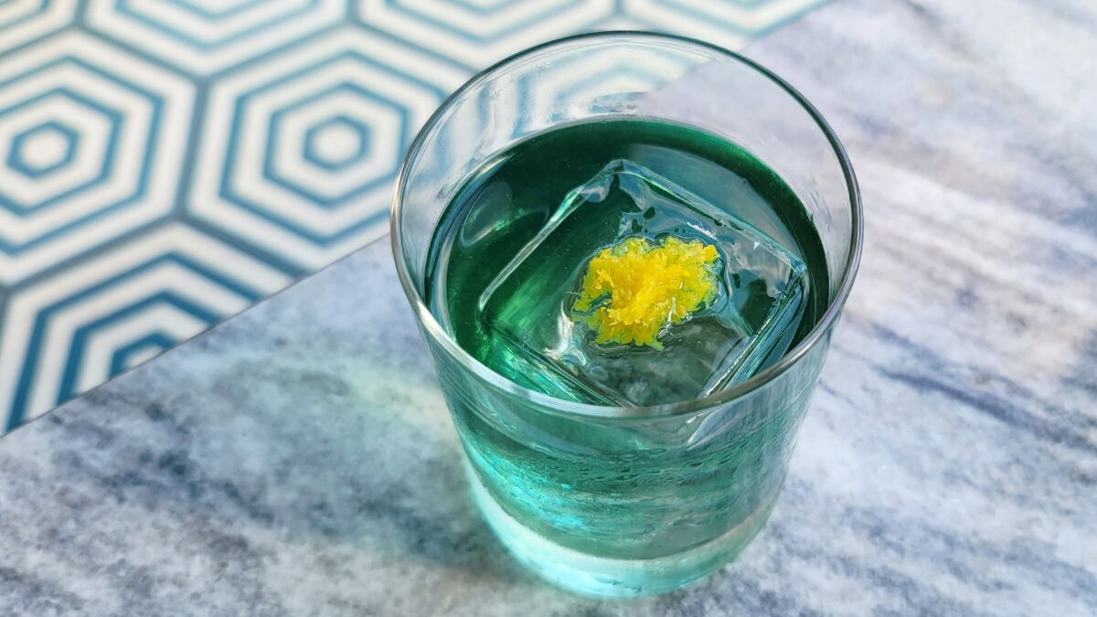 A teal-colored cocktail with an ice cube that has an edible flower inside.