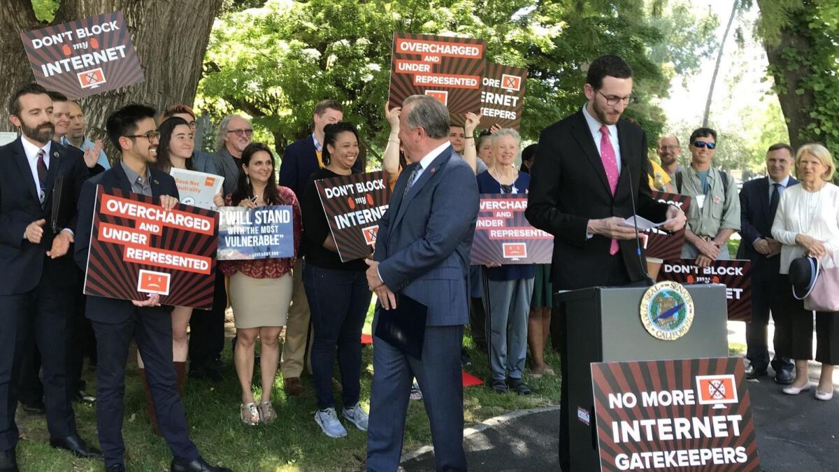 A lawmaker speaks at a lectern in front of a group of people with signs in support of net neutrality