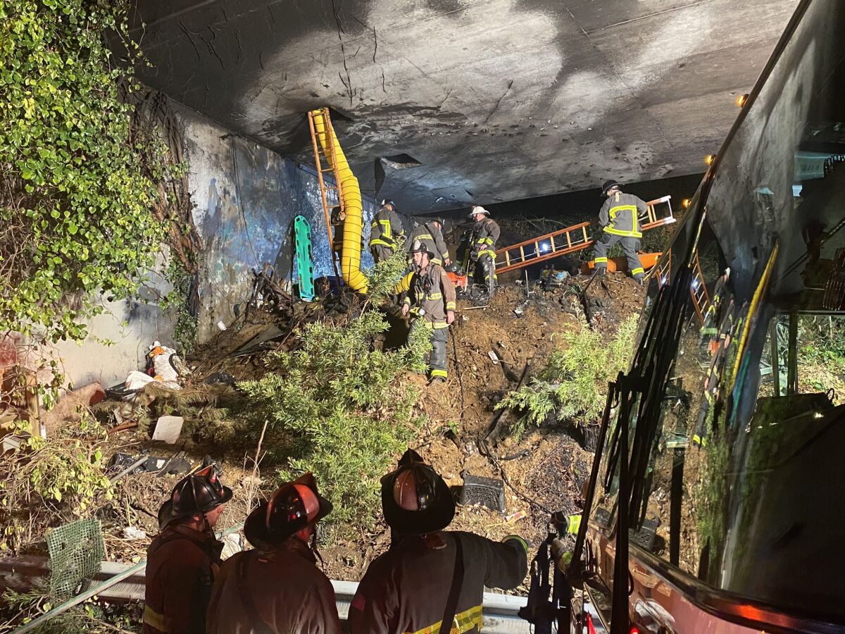 Firefighters use ladders to climb into an overpass crawlspace charred by fire above an overgrown hill