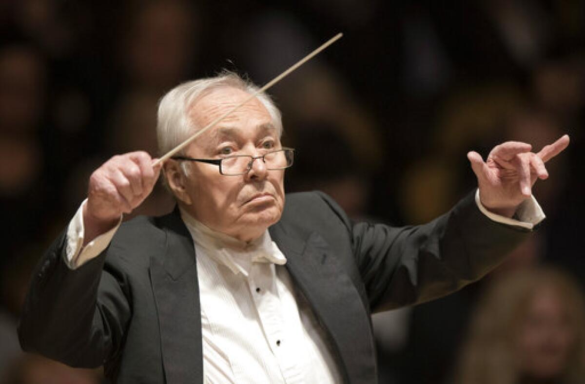 An elderly man wearing formalwear with his hands in the air conducts an orchestra 