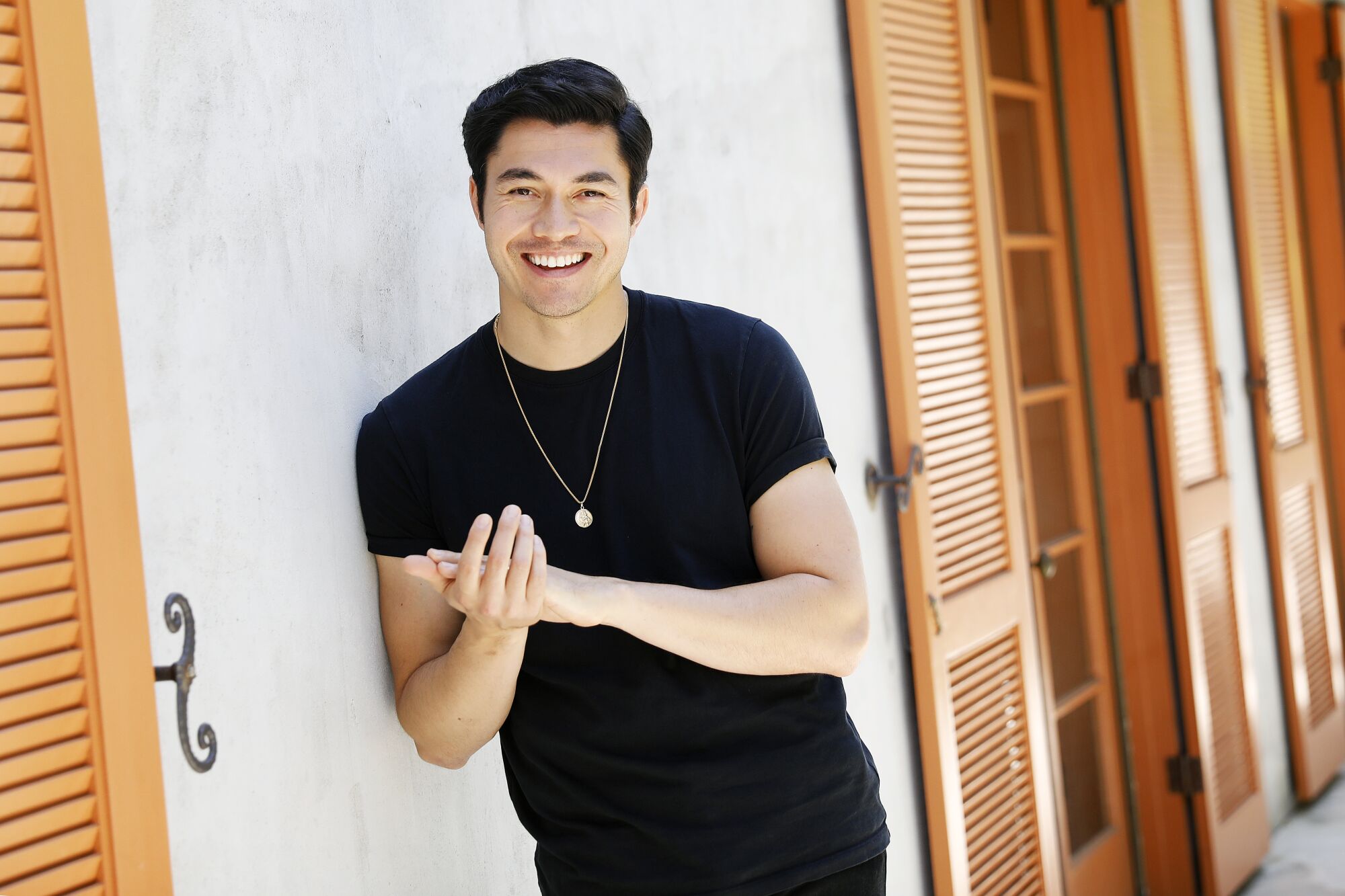 Actor Henry Golding, casually dressed in all-black, stands in front of shuttered French doors. 