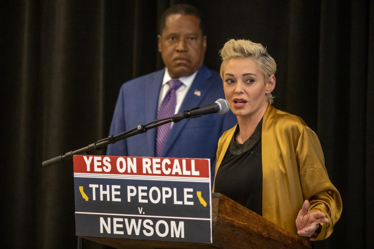 A woman in a gold silk jacket speaks at a podium while a man in a suit stands beside her