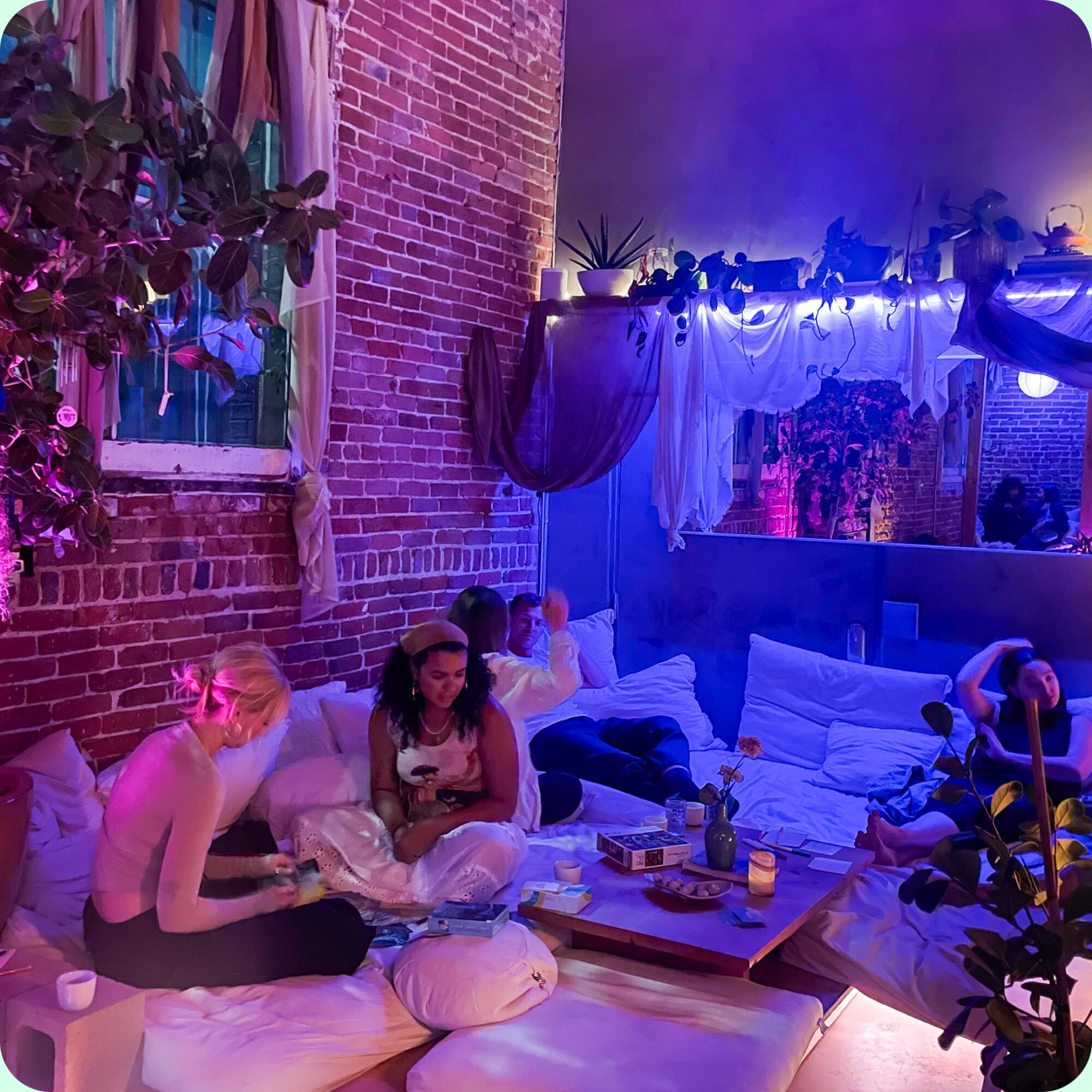 People rest in a cozy-looking room with blue lights, a brick wall, hanging plants, floor cushions and low tables.