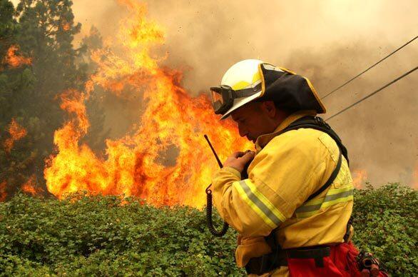 A firefighter with the Lathrop-Manteca Fire District talks on his radio as a spot fire burns through trees and brush in Concow, Calif., where firefighters continue to battle a brush fire that threatens backwoods towns and the city of Paradise.