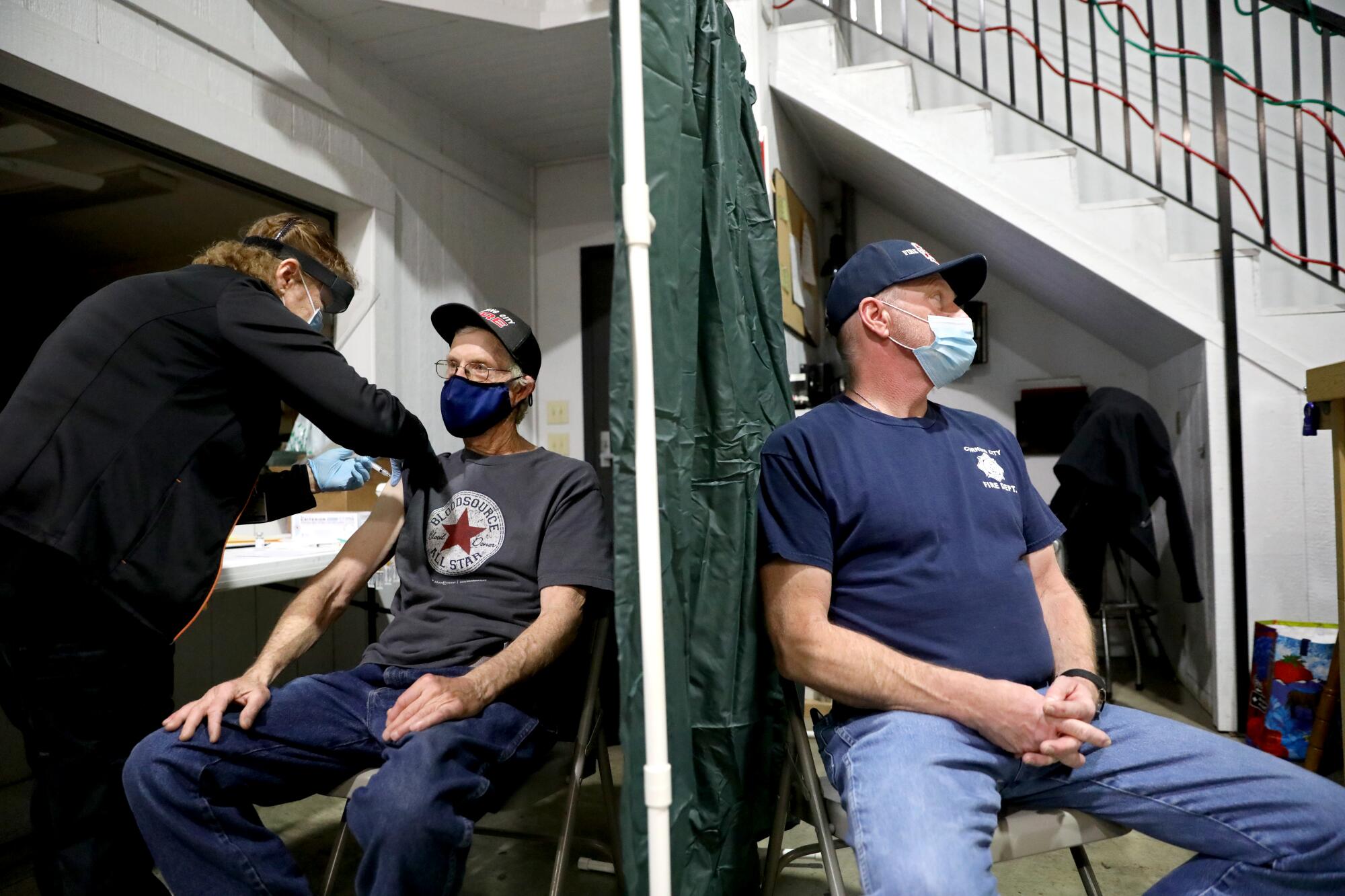 Two men sit in chairs as a women injects a vaccine shot into the arm of one