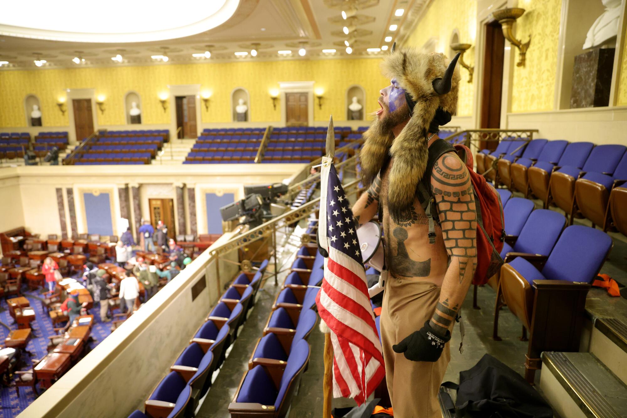 A man with tattoos and a fur headdress and flag stands, yelling, amid rows of seats in the Senate chamber.