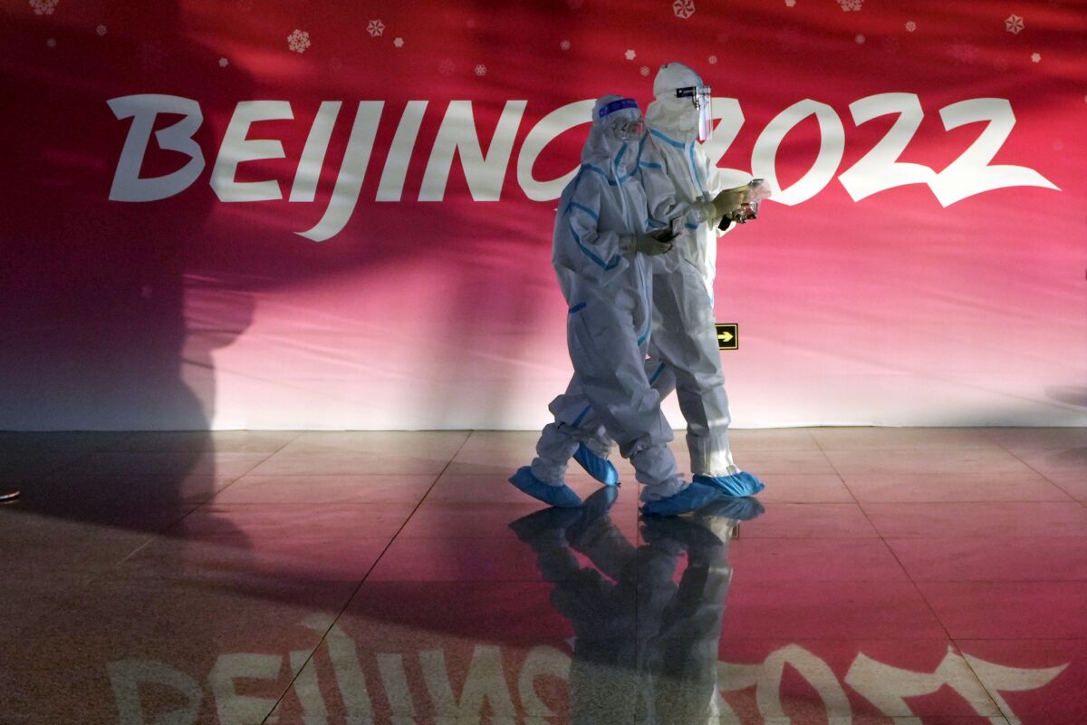 Two people in hazmat suits walk past a Beijing 2022 logo.
