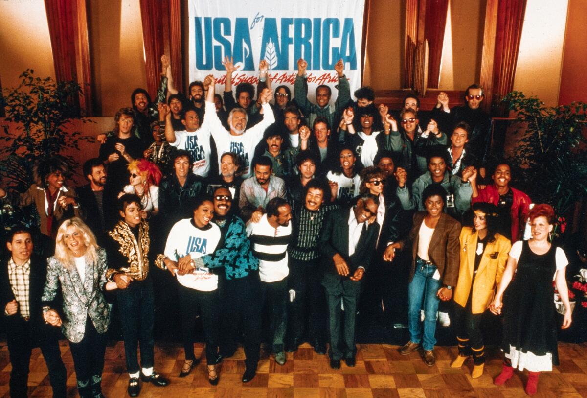 A group of musicians gathers under a sign that says USA for Africa.