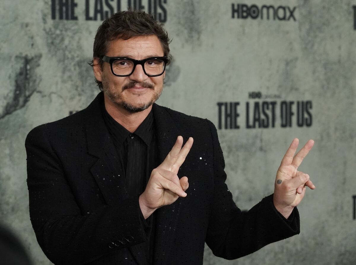 A man wearing black glasses and a black suit holds up peace signs with both hands