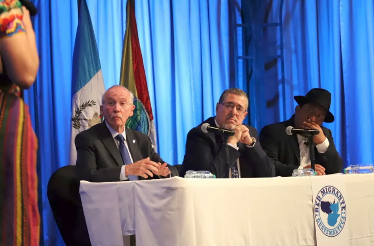 Three men in dark suits sit at a table in front of flags.