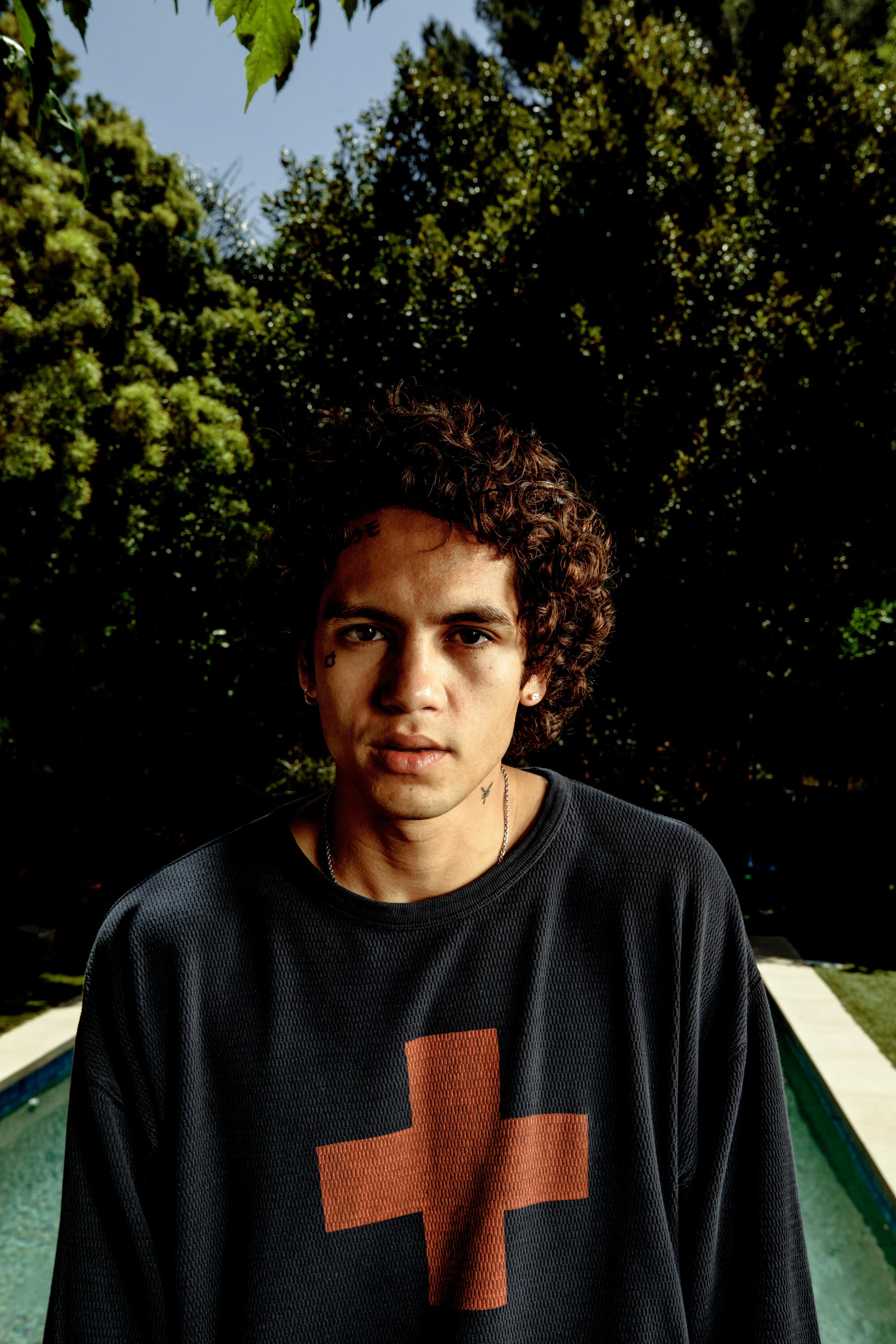 Dominic Fike in a black shirt with a red cross on it, outdoors with trees behind him