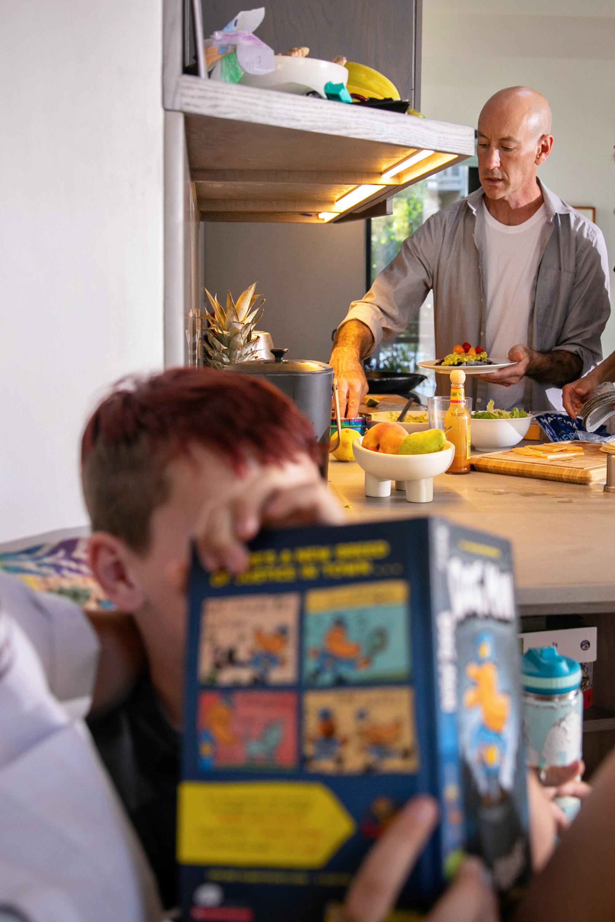 A man prepares dinner in the kitchen while a child reads in a nearby corner. 