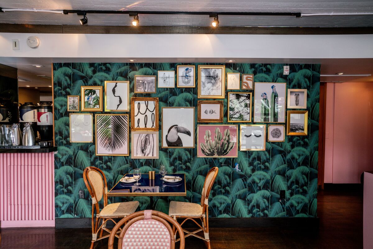 Wicker chairs at a table in a restaurant with green leafy wallpaper on a wall hung with lots of framed art.