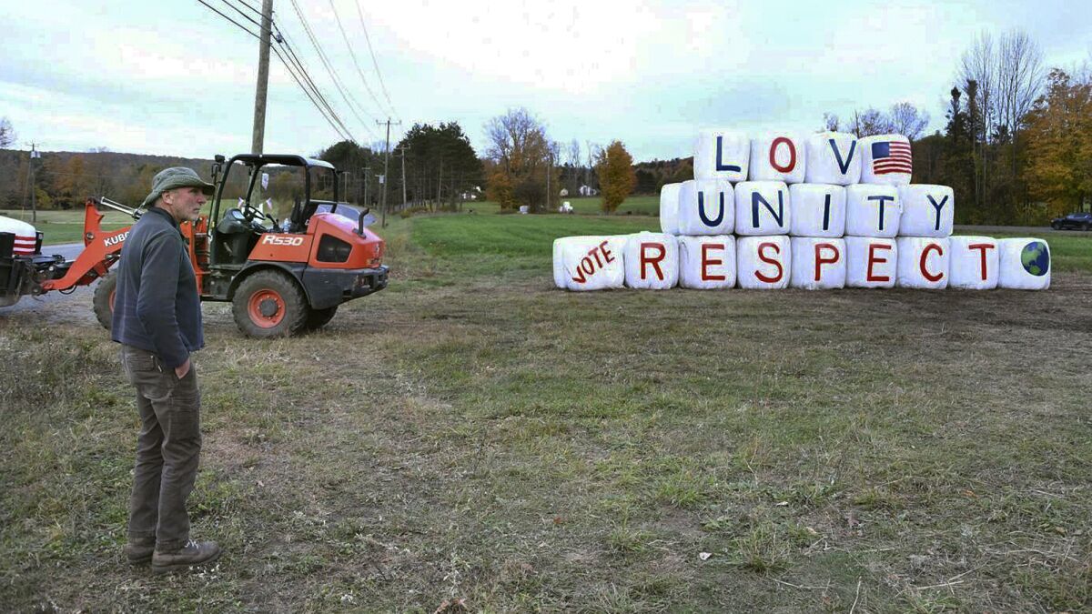 Farmer Dicken Crane stands beside hay bales painted with the words "Love, Unity, Respect."
